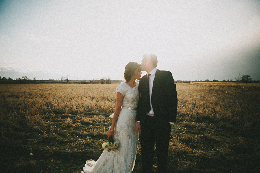 Film-style photo of a groom kissing his bride on the forehead in an open field