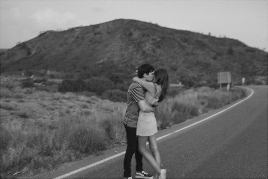 Black and white film photo of a couple kissing in the middle of the desert road.